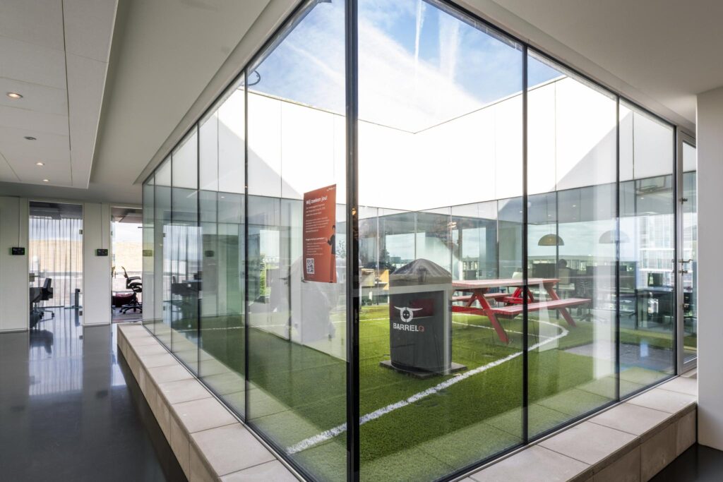 Glass-walled indoor courtyard at Krijn Taconiskade featuring artificial grass, a red picnic table, and a BarrelQ barbecue.